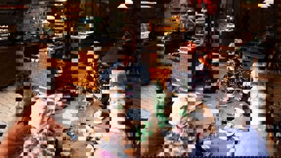 A group of six people sit around a round wooden table in a busy, rustic-industrial Outlier.   They are drinking coffee and talking, with an open kitchen visible in the background.