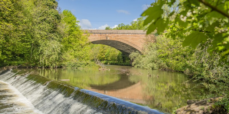 A wide stone bridge crosses a river surrounded by green trees. The river flows into a mini waterfall.