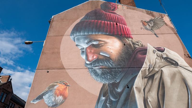 A large outdoor mural on the side of a building, depicting a bearded man in a red hat with two robins.