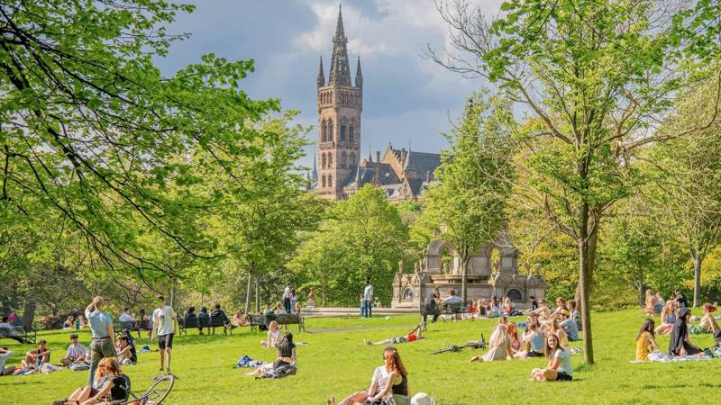 People relax on a sunny day in a green park with a grand, spired building visible in the background.