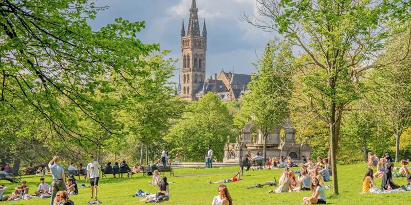 People relax on a sunny day in a green park with a grand, spired building visible in the background.
