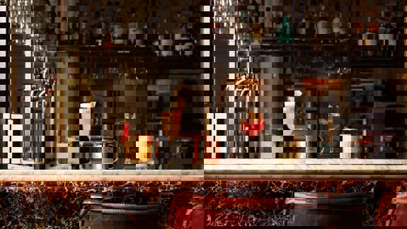 Five distinct cocktails sit on a marble-topped bar with brass fittings, set against a backdrop of neatly stacked bottles and barware.