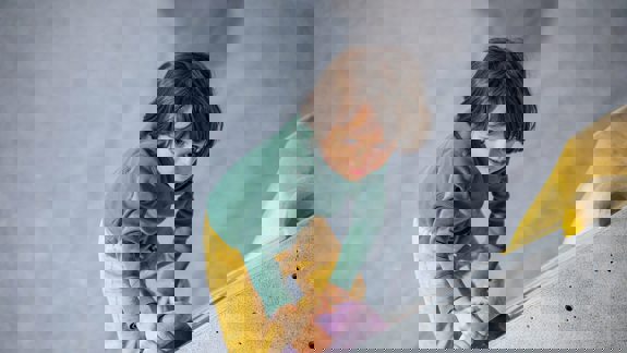 A young child in a green shirt and yellow trousers indoor climbing, gripping a purple handhold on a grey climbing wall.