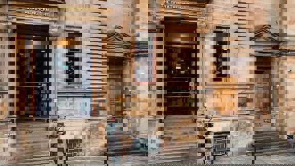 A man in a kilt plays bagpipes on the steps of a building with a sign that says "The National Piping Centre" above the entrance.