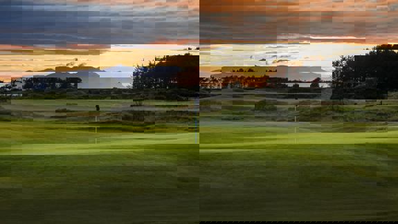 Sunset over Dundonald Links golf course with rolling fairways, flagstick, and the mountains silhouetted in the distance.