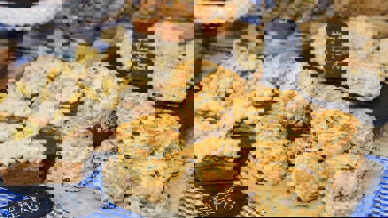 A close-up shows a display of baked goods, including a pile of golden raisin scones on a wooden board, banana  and peanut butter loaves, and vegan apple and cinnamon scones, all artfully arranged for sale.