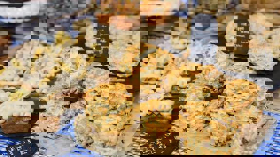 A close-up shows a display of baked goods, including a pile of golden raisin scones on a wooden board, banana and peanut butter loaves, and vegan apple and cinnamon scones, all artfully arranged for sale.