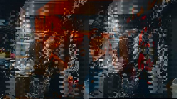 A person wearing an apron smiles while stirring a pot in a bustling kitchen with shelves and hanging utensils.