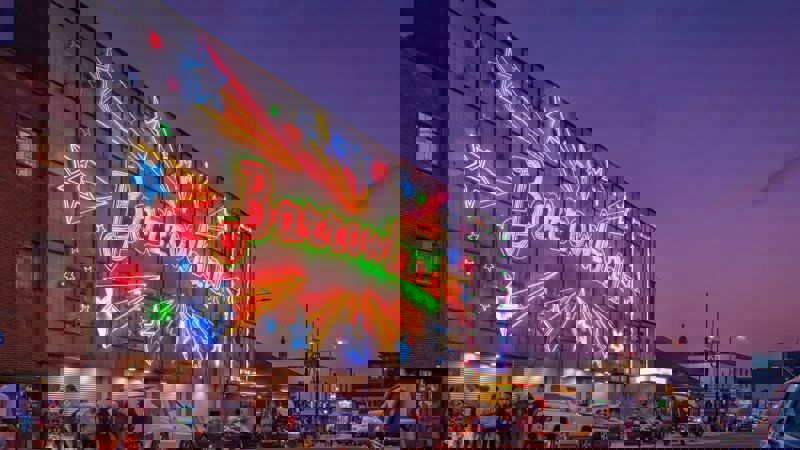 The famous Barrowland sign which says the word Barrowland surrounded by shooting stars is lit up against a dusk sky. People mingle at the front of the venue.