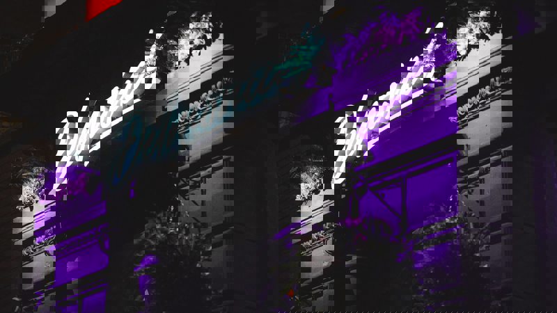 Night-time view of a building exterior with a bright, cursive, neon "Delmonica's" sign over the entrance, illuminated by vibrant purple light.