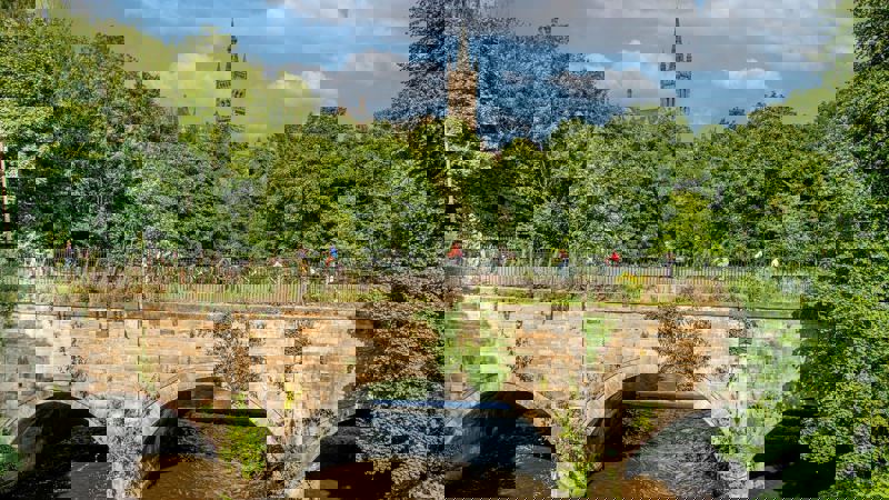 A group of cyclists riding across a stone bridge over a river, with trees and a distant tower visible behind them.