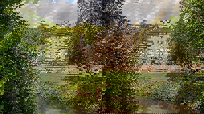 A grand manor house overlooking a river and lush green trees in Pollok Park, Glasgow.