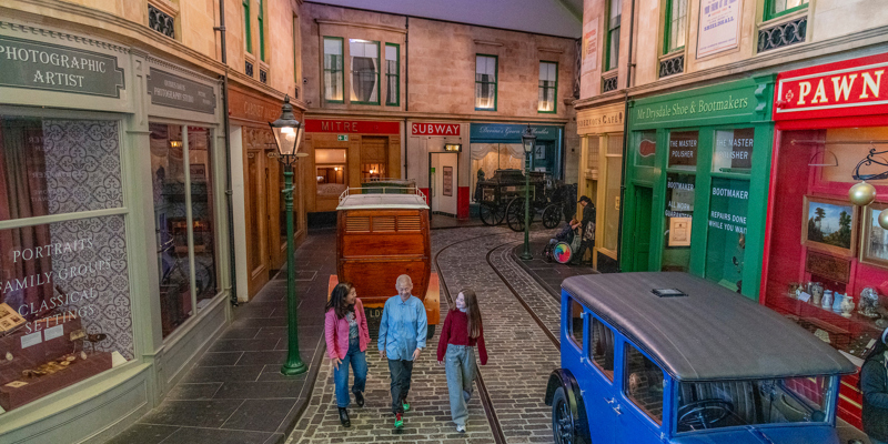 A cobbled indoor street scene with vintage shopfronts and early 20th‑century vehicles on display, as three people walk beneath string lights in a museum gallery styled like an old town.