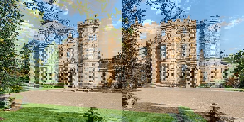 A grand stone castle with a paved courtyard and green lawn is shown on a sunny day with a blue sky and a few clouds.
