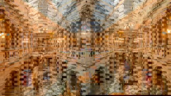 Grand interior of Kelvingrove Art Gallery and Museum. A Spitfire plane hangs from the ceiling over taxidermy animals, with ornate balconies and a glass roof above.