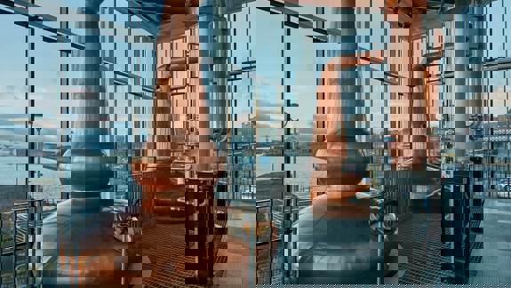 Copper whisky stills at Clydeside Distillery in Glasgow, with the River Clyde and a tall ship visible outside the large window