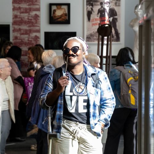 A person in sunglasses and a "LOVE" T-shirt smiles while holding a fake mustache on a stick. The background shows a lively, crowded room with people and art on the walls, conveying a cheerful and playful atmosphere.