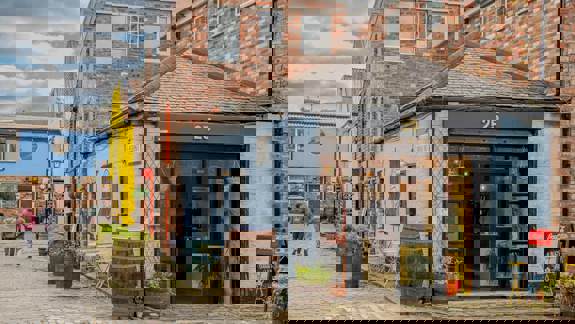A street view of a small, light-blue cafe with outdoor seating and plants in whiskey barrels. Other brightly colored brick buildings line the cobblestone alleyway in Glasgow's Hidden Lane.
