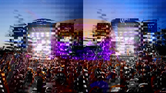 A massive crowd with raised hands faces a brightly lit TRNSMT main stage at twilight, illuminated by vivid purple lights and screen displays.