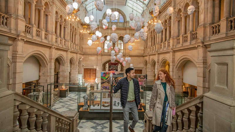 Two people descend a grand stone staircase inside a museum, beneath an art installation of many white, floating heads suspended in a large, arched hall.