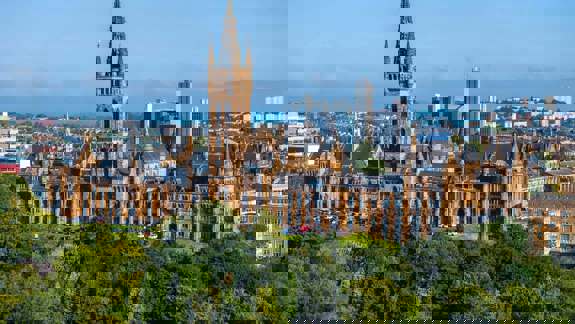 Panoramic view of the historic University of Glasgow building, with trees in the foreground and city skyline behind.