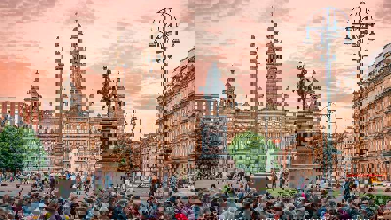 George Square in Glasgow at sunset, with a crowd of people gathered in the foreground and the illuminated City Chambers building and statues in the background.