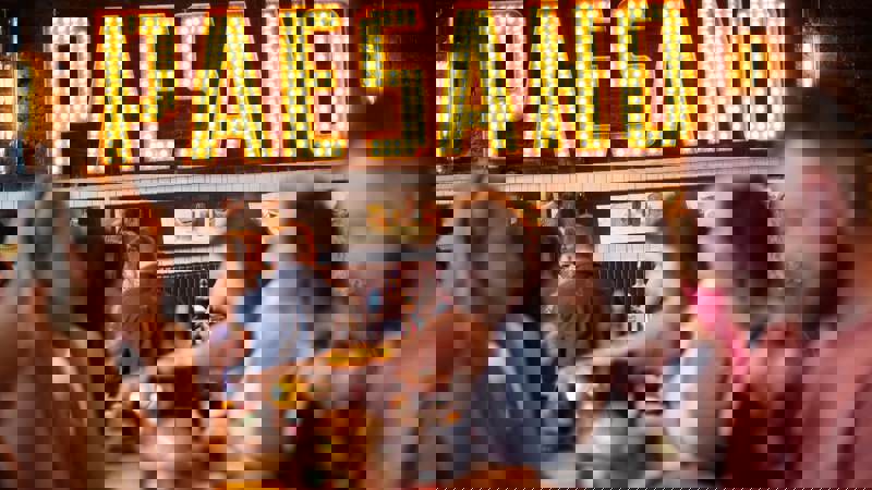 A group of people sitting at tables in a restaurant, with a large, lighted sign that reads "PAESANO" in the background.