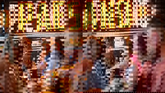 A group of people sitting at tables in a restaurant, with a large, lighted sign that reads "PAESANO" in the background.