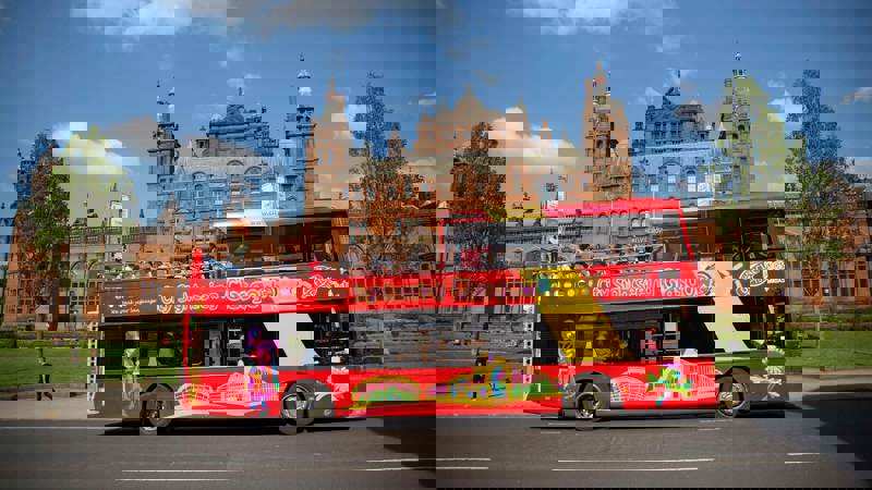 A double-decker bus with "City Sightseeing Glasgow" written on its side, driving past a large, red brick building.