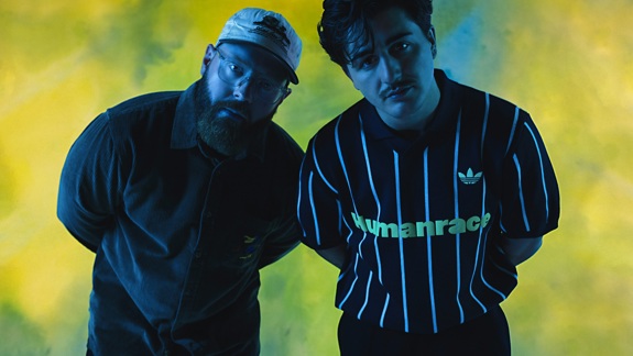 Two men posing against a blue and yellow background, leaning slightly forward toward the camera under studio lighting.