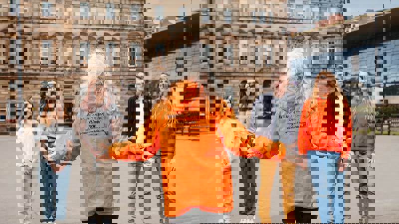A woman in an orange "Walking Tours in Glasgow" jacket with outstretched arms addresses a small group of people in a city square, with historic buildings in the background.