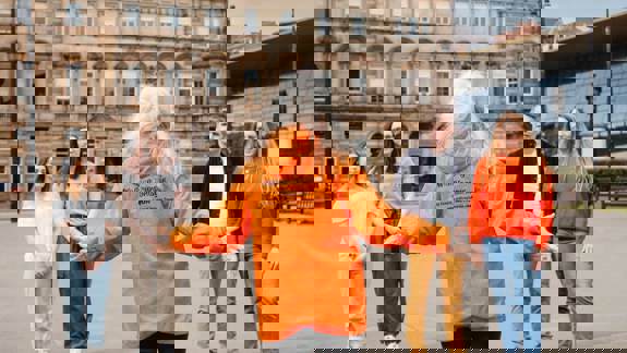 A woman in an orange "Walking Tours in Glasgow" jacket with outstretched arms addresses a small group of people in a city square, with historic buildings in the background.
