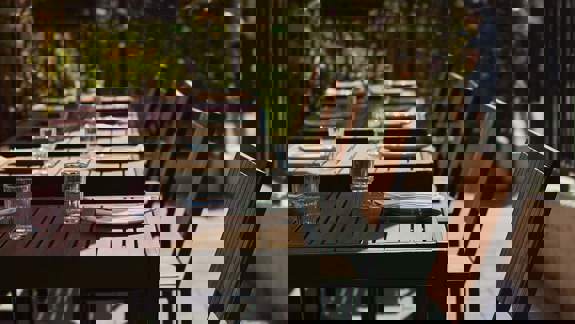 Outdoor dining area with a wooden table set with glasses and plates, surrounded by chairs under a red umbrella.