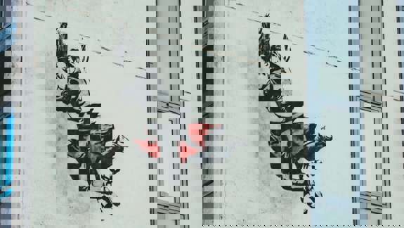 A stencil-style mural on a white building wall by The Rebel Bear, depicting two figures locked in a romantic embrace while appearing to fall or float through the air.