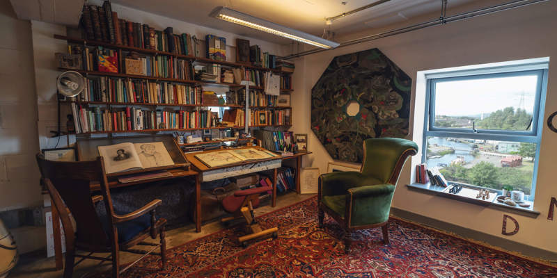 A room with a red patterned rug, a large bookcase filled with books, and a desk with drawing supplies. In the center is a green velvet armchair.
