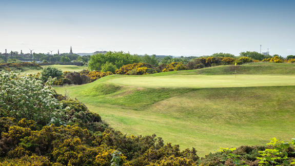 Elevated green at Irvine Golf Club surrounded by gorse bushes, with views across the town and wind turbines in the distance.
