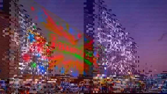 The vibrant, multi-colored neon sign of the Barrowland Ballroom, adorned with stars, illuminates a city street at dusk as people gather outside.
