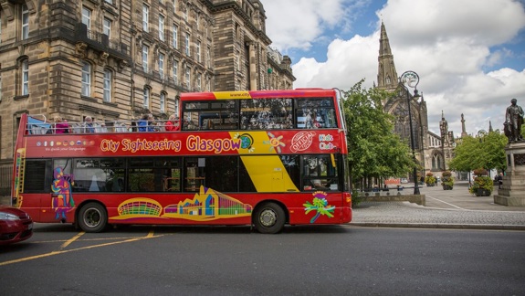 A red and yellow Glasgow sightseeing bus with people on the top deck is stopped on a street in front of a large, historic stone building and a church with a spire.