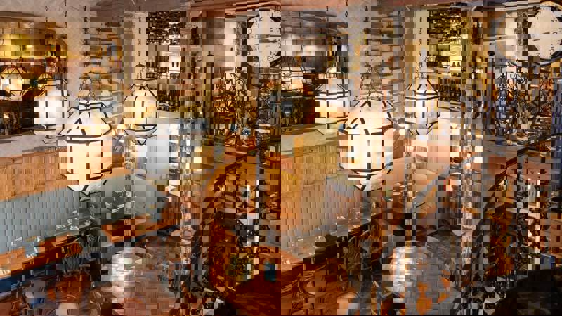 The rustic, two-story interior of a restaurant featuring wooden tables, large hanging lights, and a spiral staircase.