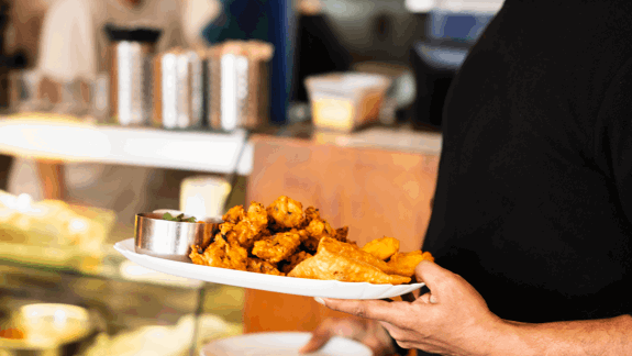 White plate with fried pakoras, samosas, and dipping sauce held by person in black shirt, with kitchen counter and utensils in background.