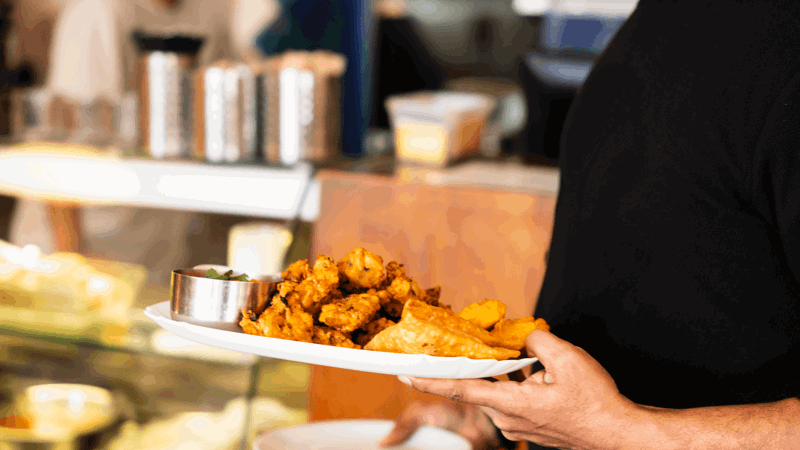 White plate with fried pakoras, samosas, and dipping sauce held by person in black shirt, with kitchen counter and utensils in background.