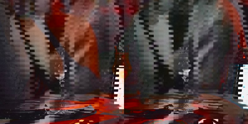 A delicate stem glass holds a pale gold drink on a dimly lit bar, with blurred figures and warm red lighting in the background.