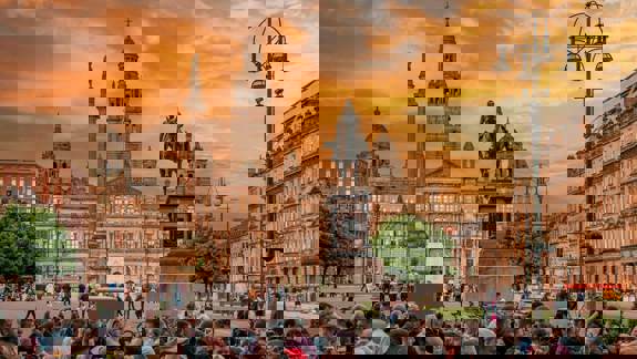 A vibrant photo of a crowd of people gathered in Glasgow's George Square at sunset. In the background, the Glasgow City Chambers stands under a beautiful orange and cloudy sky.