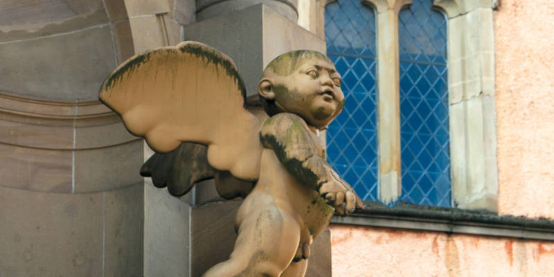 A weathered stone sculpture of a plump cherub with wings clinging to a building's exterior.