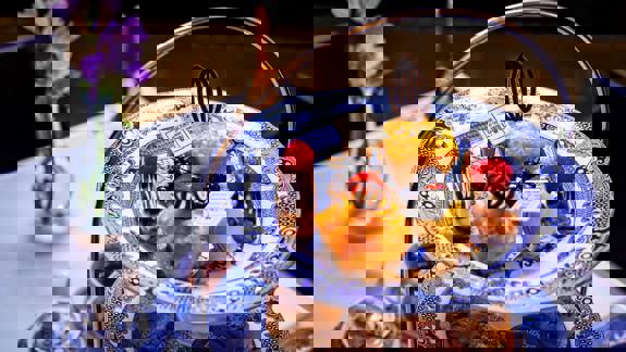 Close-up of delicate pastries, including muffins and chocolate-decorated eclairs, on a blue and white patterned plate with a "Mackintosh at The Willow" white chocolate tag.