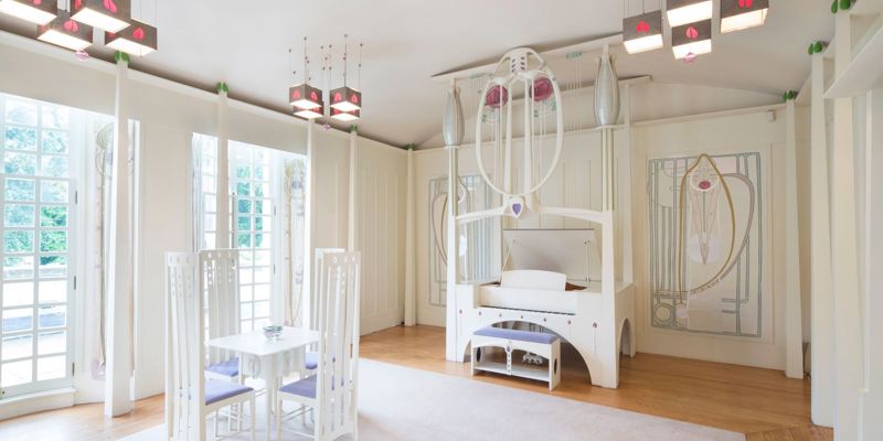 A white-painted room with furniture and light fixtures in the Mackintosh style, including a piano and a small dining table.
