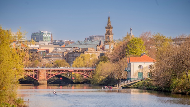 View of the River Clyde in Glasgow on a sunny day, showing rowers on the water, a red bridge, and city buildings with towers and trees in the background.