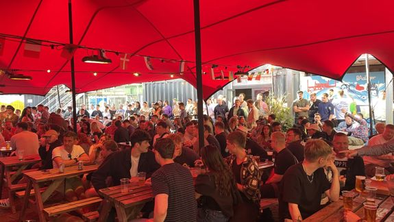 Large crowd seated at wooden picnic tables under a bright red canopy in an outdoor venue, with drinks on the tables and more people standing in the background