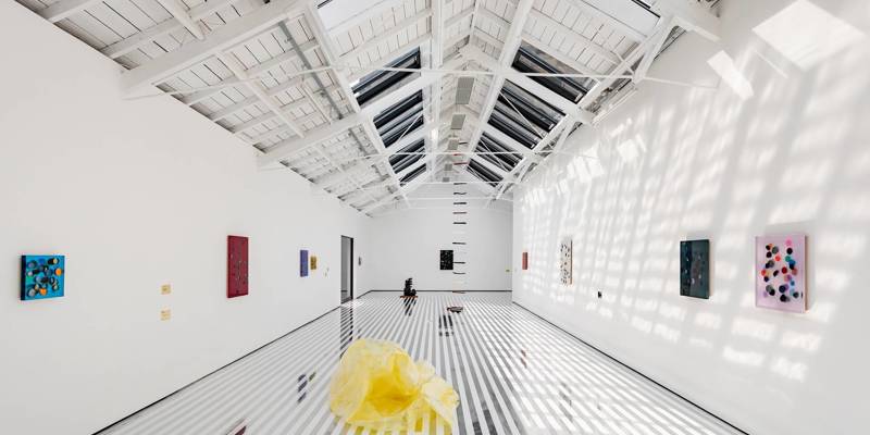 A bright, white-walled art gallery with a striped floor, several small paintings, a central yellow sculpture, and a sloped, skylight ceiling.