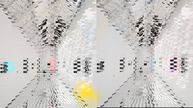 A bright, white-walled art gallery with a striped floor, several small paintings, a central yellow sculpture, and a sloped, skylight ceiling.